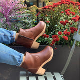 A pair of tan vegan leather clog booties on a woman's feet. Ankle height shaft with pull tab in back. Inside zipper closure. Blonde wooden sole. Staples around outsole to connect material to sole. Flowers in background.